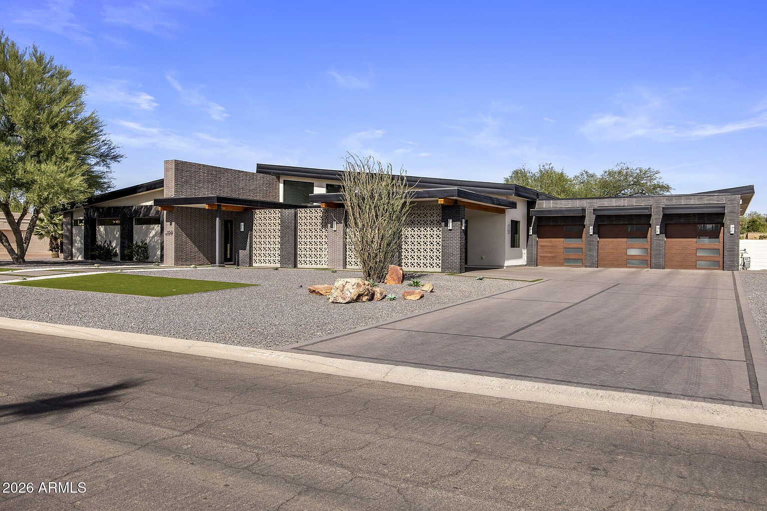 Full daytime facade: long mid-century roofline, breeze-block panels between brick piers, three-car garage with reclaimed-wood doors, and an ocotillo cactus marking the entry.