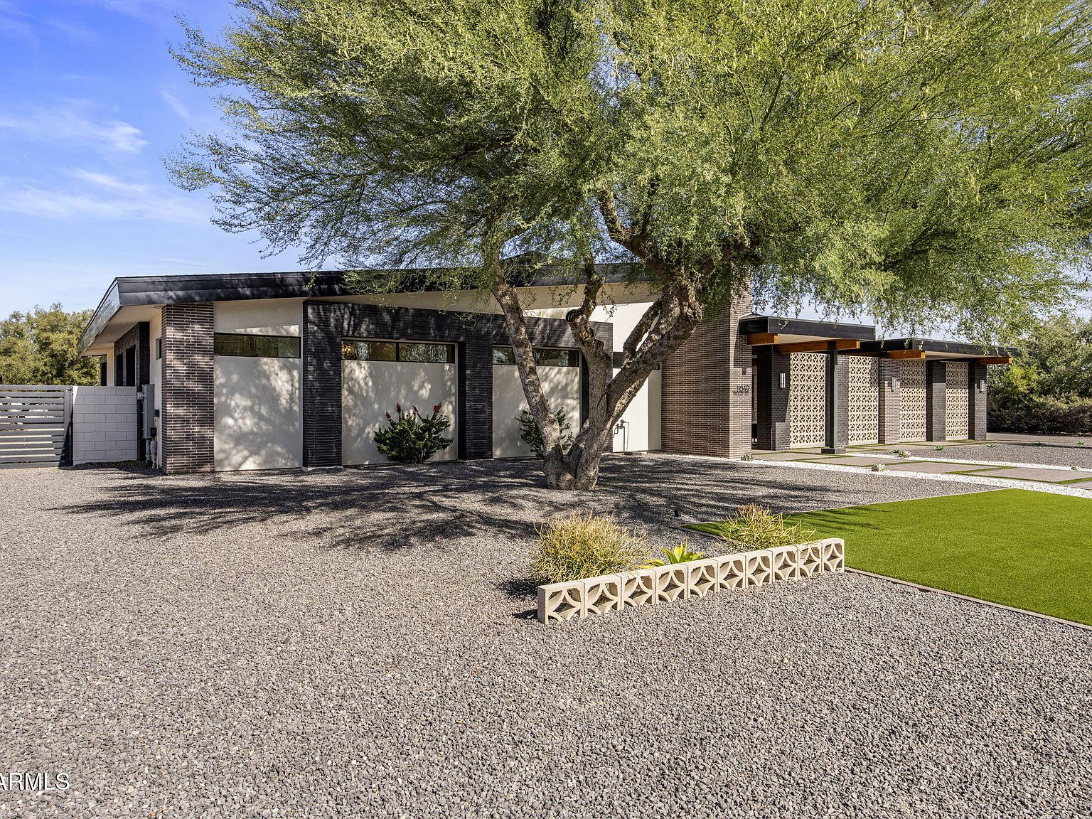 Wide approach from the driveway at 11649 N Miller Road: the breeze-block facade in full daylight, side garage doors, decomposed-granite grounds, and a mature mesquite tree framing the entry.
