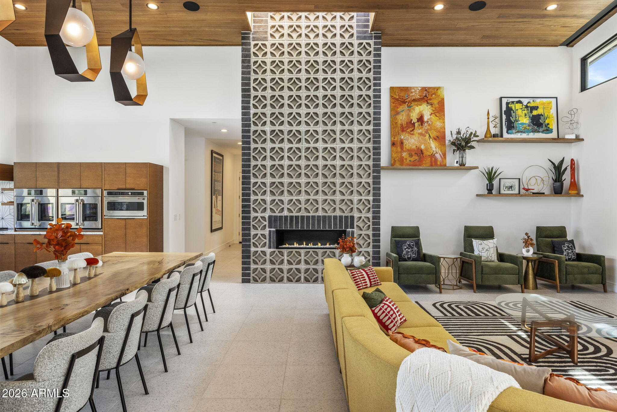 The great room: double-height ceilings clad in warm wood, a full-height breeze-block fireplace wall, long communal dining counter, and seating arranged around a low brick hearth.