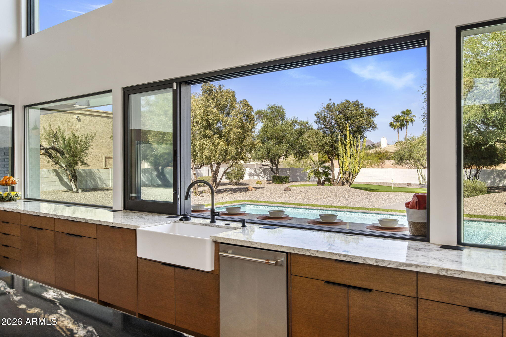 The kitchen window at the sink: walnut cabinetry, a marble counter, an apron-front basin, and a long picture window framing the pool and the desert garden beyond.
