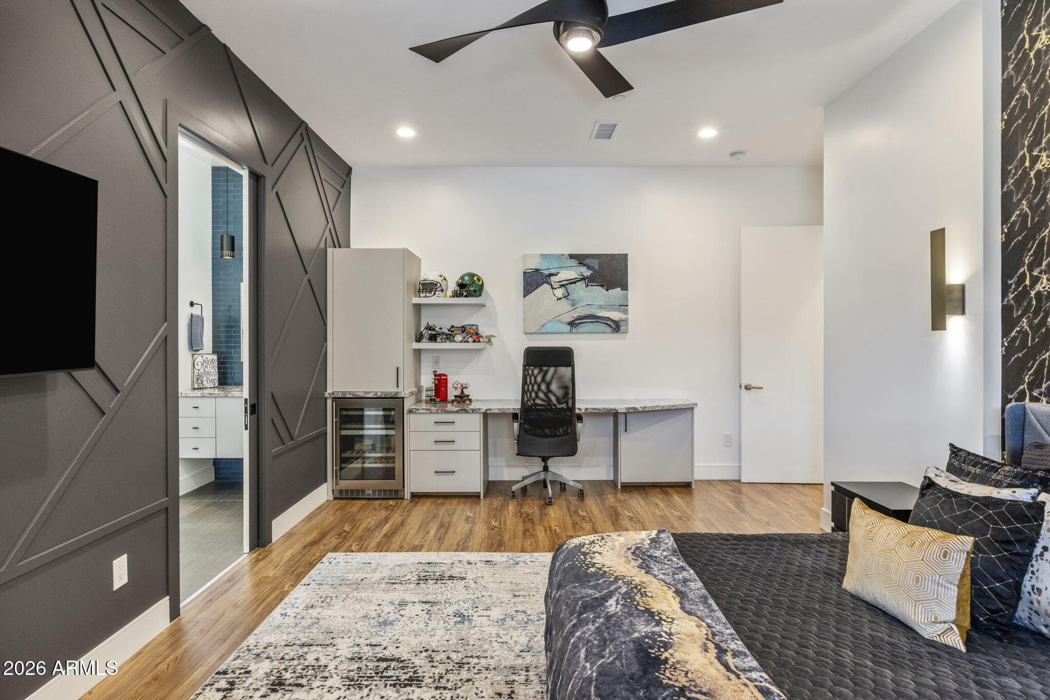 A bedroom suite with a charcoal paneled accent wall, custom built-in desk and beverage cooler, modern art, and oak flooring catching afternoon light.