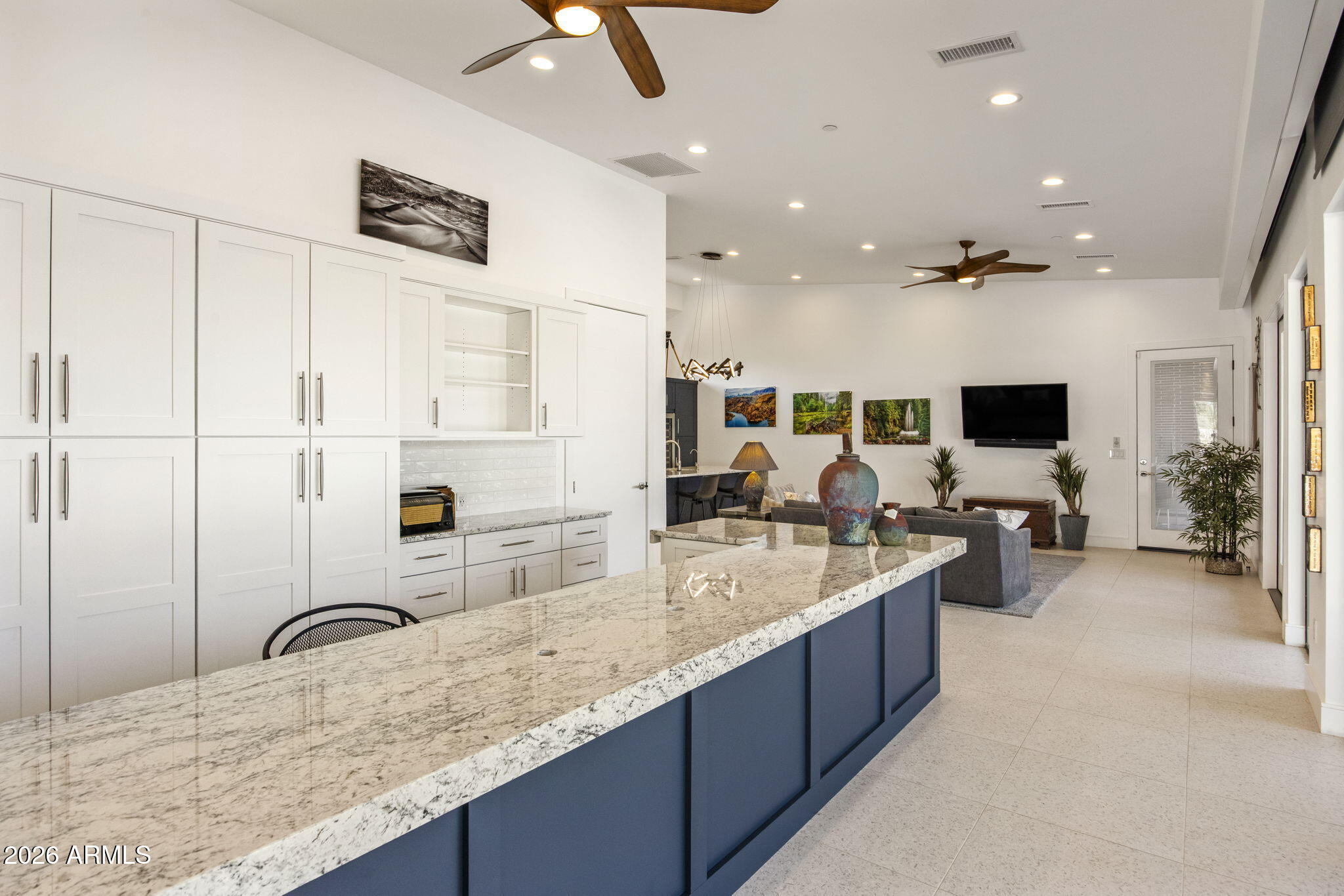 The kitchen and great room looking east: an oversized navy island under a sculptural fan, white cabinetry, terrazzo flooring, and the living room opening past the dining table.