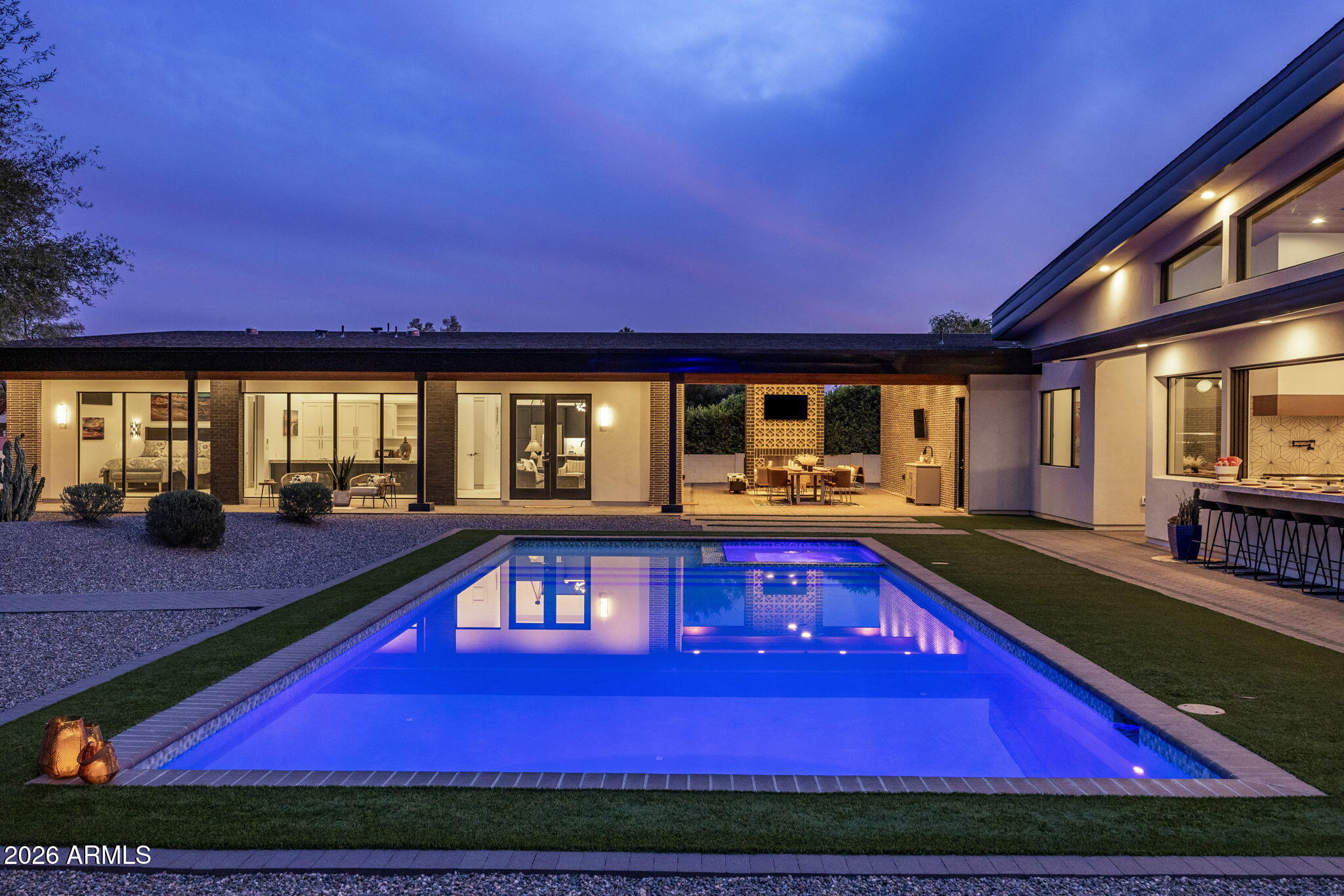 The backyard at twilight: a long rectilinear pool lit cobalt blue, lounge seating under the covered terrace, and the rear of the house glowing through floor-to-ceiling glass.
