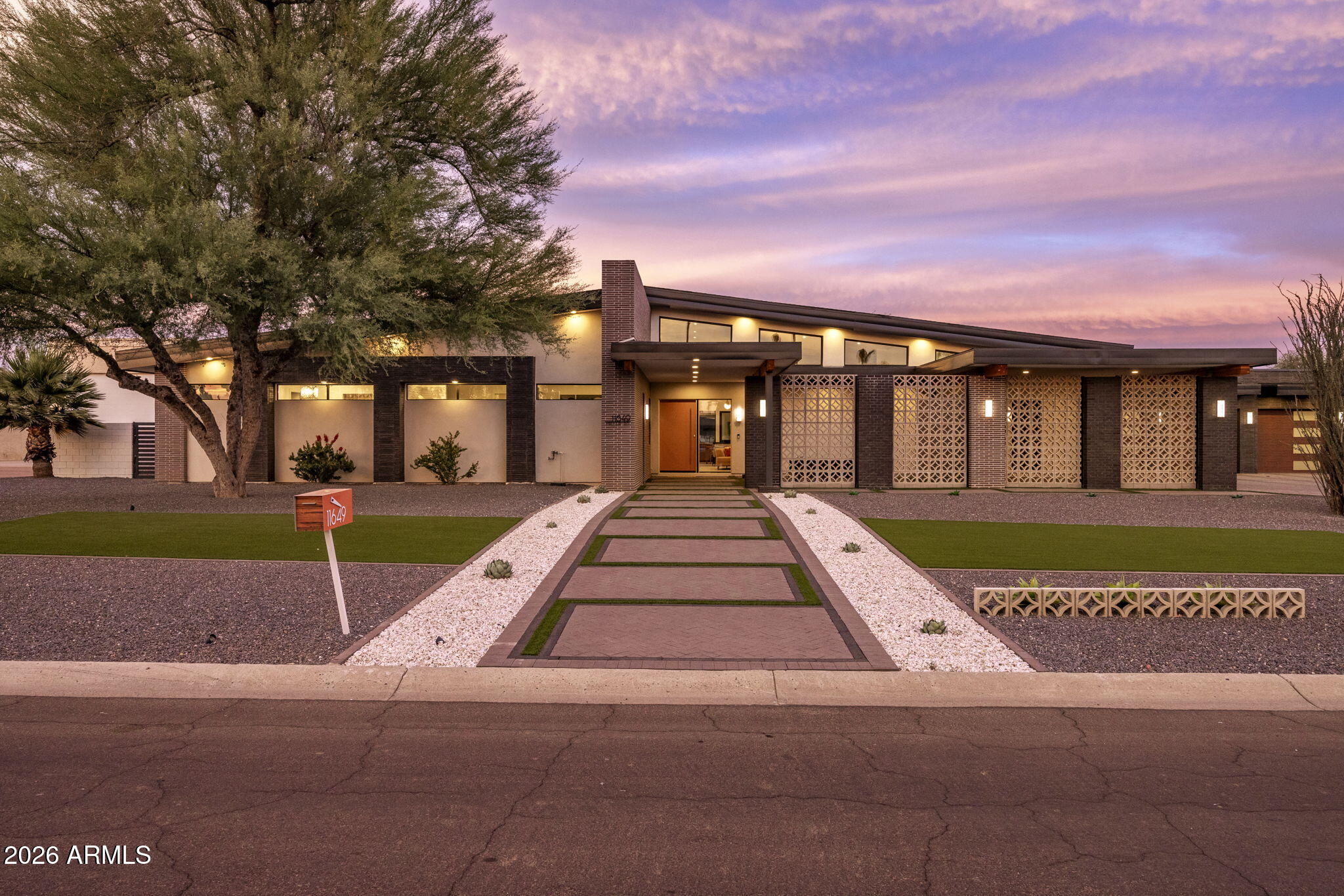 The front elevation at sunset: low mid-century rooflines lit warm, breeze-block panels in silhouette, a clean paver walkway, and a pink-and-violet desert sky overhead.